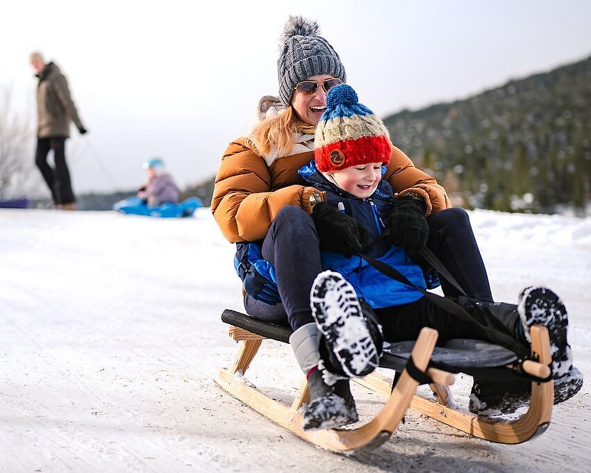 Mutter und Kind rodeln auf einem Holzschlitten einen verschneiten Hang hinunter.