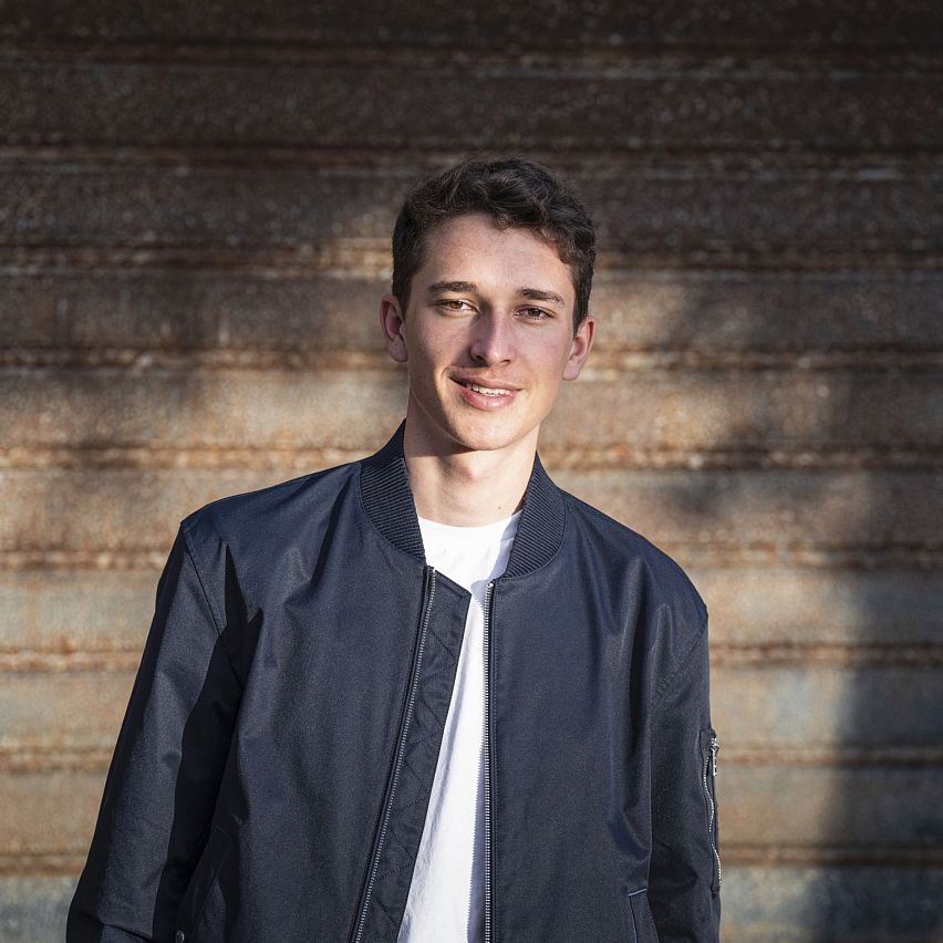 Young man in dark jacket stands against a textured wall, smiling.