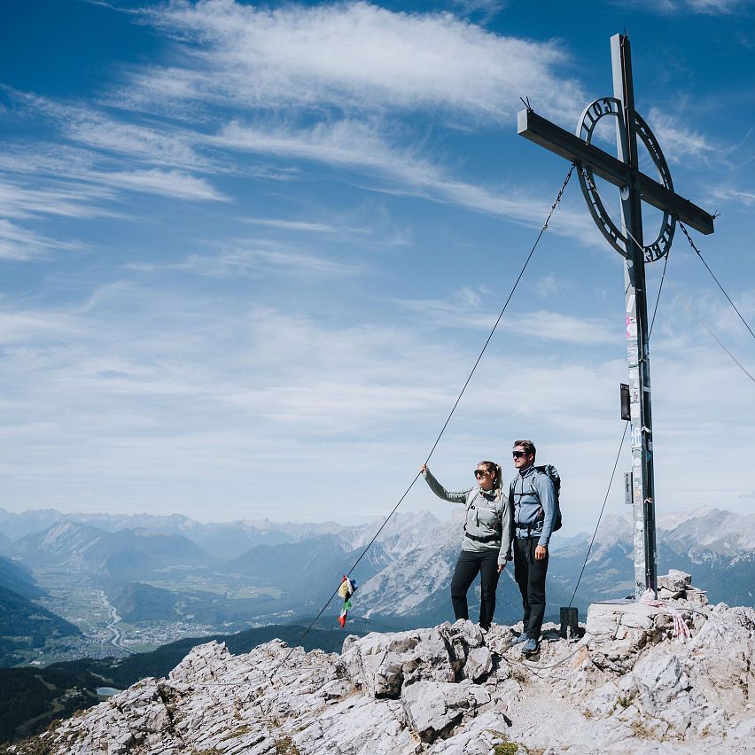 A couple at a mountain peak beside a large cross with panoramic views in the background.