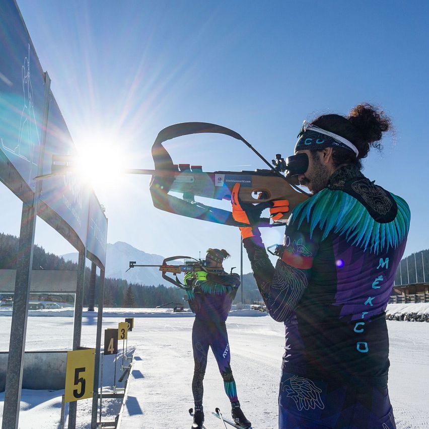 Zwei Sportler in bunten Anzügen schießen auf eine Zielscheibe bei sonnigem Wetter.