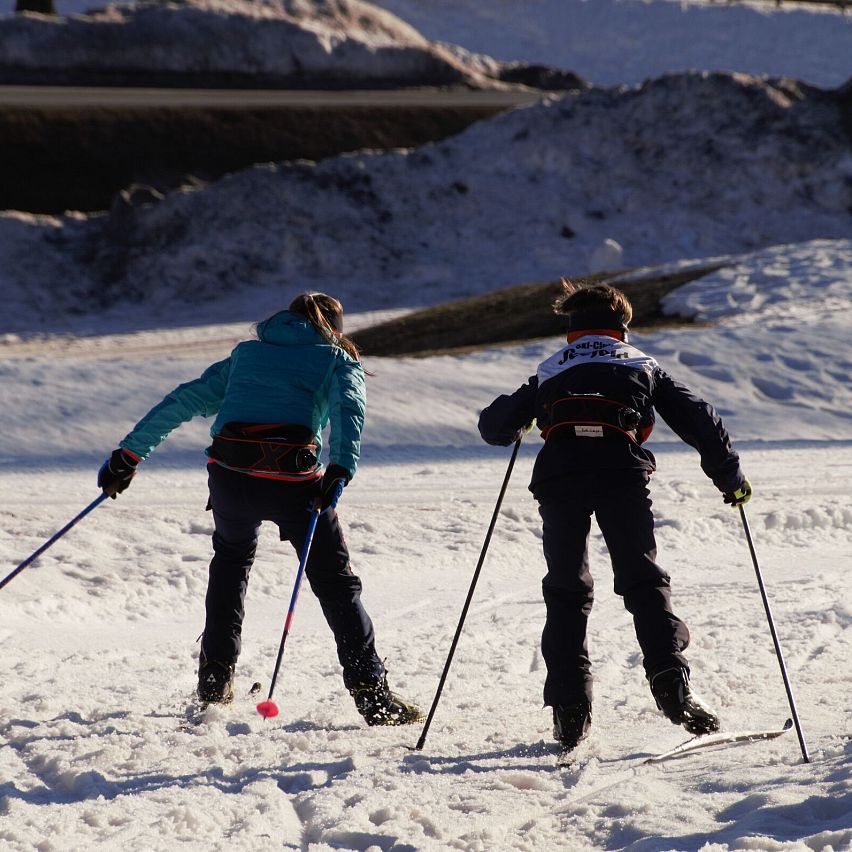 Zwei Kinder beim Langlaufen im Schnee mit Skistöcken, sonnige Winterlandschaft.