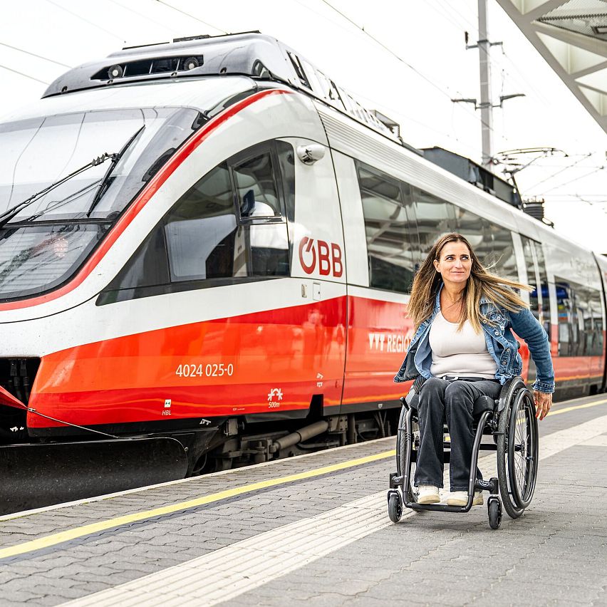 Frau im Rollstuhl an Bahnsteig mit einfahrendem ÖBB-Zug im Hintergrund.