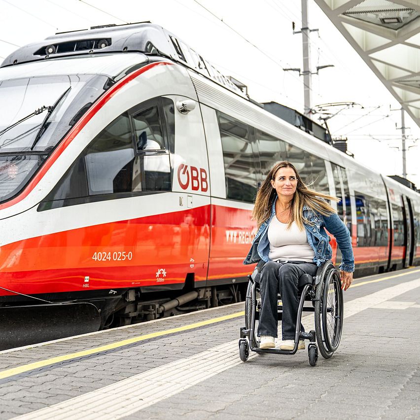 Frau im Rollstuhl an Bahnsteig mit einfahrendem ÖBB-Zug im Hintergrund.