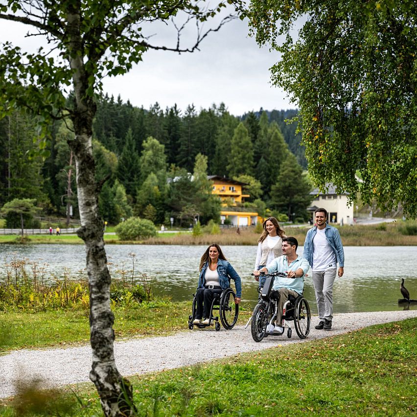 Familie am Seeweg in der Region Seefeld, zwei Personen in Rollstühlen, malerische Umgebung mit Bäumen und bewölktem Himmel.