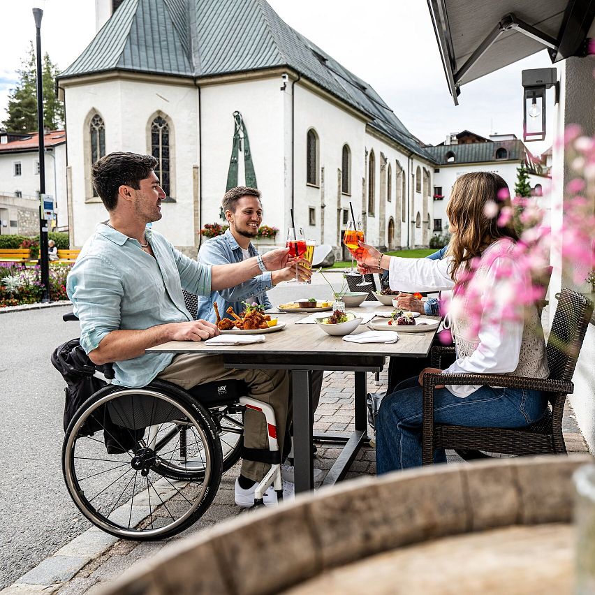 Drei Personen essen draußen in einem Bergdorf Nähe der Region Seefeld, mit einer Kirche im Hintergrund. Eine Person sitzt im Rollstuhl.