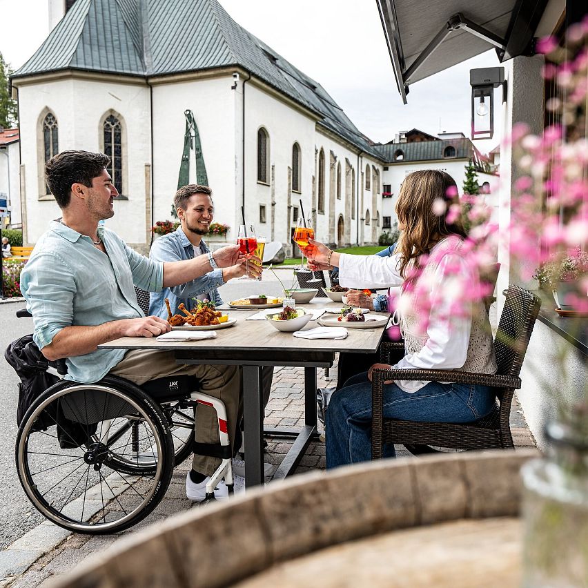 Drei Personen essen draußen in einem Bergdorf Nähe der Region Seefeld, mit einer Kirche im Hintergrund. Eine Person sitzt im Rollstuhl.