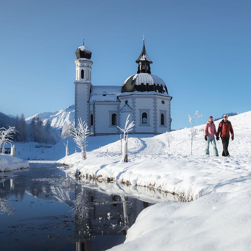 Verschneite Kirche in Winterlandschaft, Wanderer im Vordergrund. Region Seefeld. Klarer Himmel, blau.