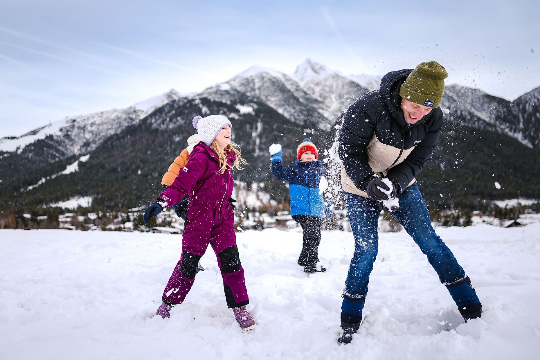 Drei Personen im Schnee vor Bergen in der Region Seefeld. Kinder werfen Schnee in die Luft. Winterkleidung, blauer Himmel.