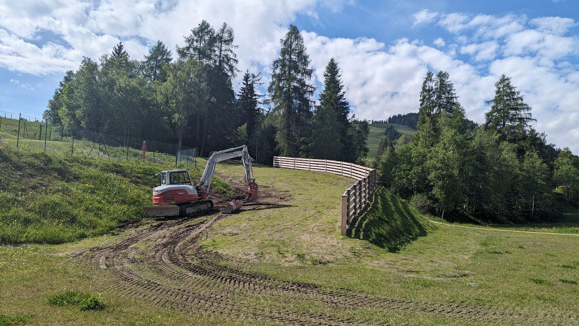 A construction machine operating on a grassy slope next to a wooden fence.
