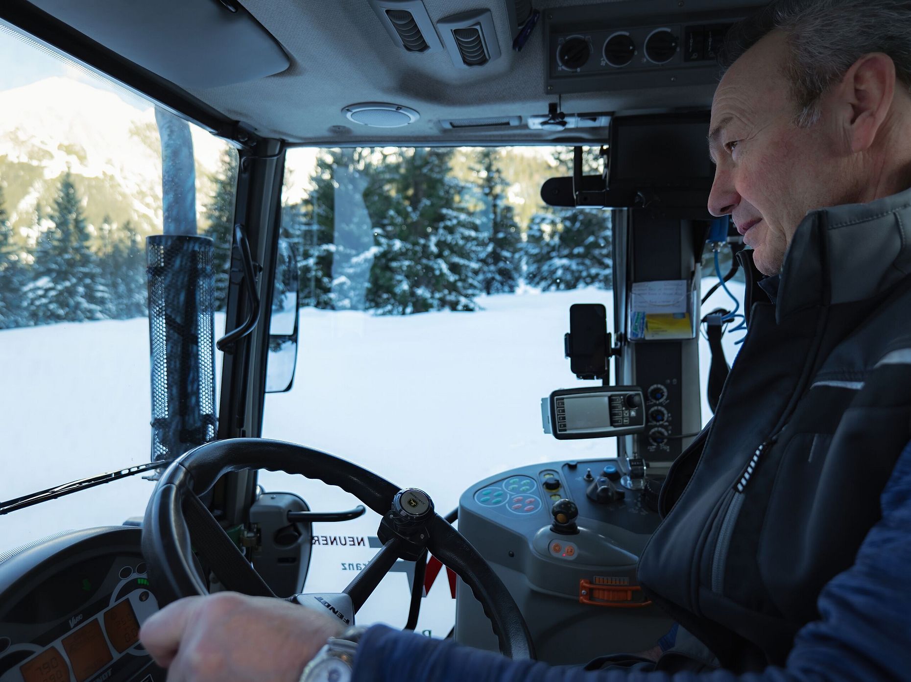 Mann fährt Schneeraupe durch verschneite Winterlandschaft, Berge im Hintergrund.