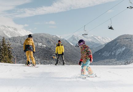 Ein Kind fährt Ski, während zwei Erwachsene zuschauen, umgeben von schneebedeckten Bergen.