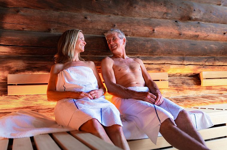 A man and woman in towels sitting in a wooden sauna, engaging in a friendly conversation.