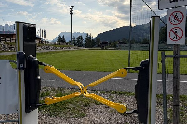 Zugang mit Drehkreuz an einem Fußballplatz, im Hintergrund Berge und grüner Rasen.