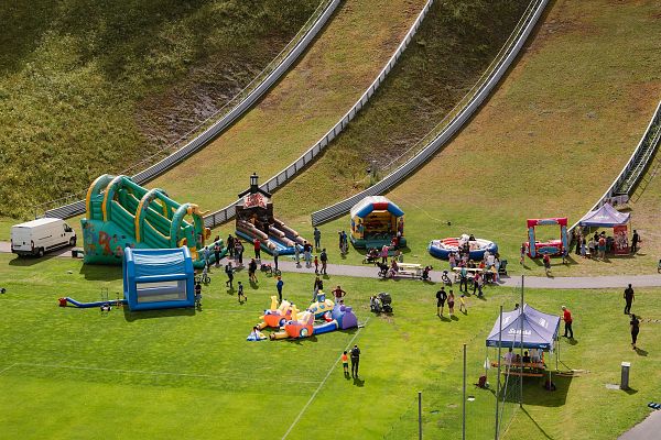 Eine Wiese mit mehreren Hüpfburgen, Spielständen und Menschen, die an einem Freizeitevent teilnehmen.