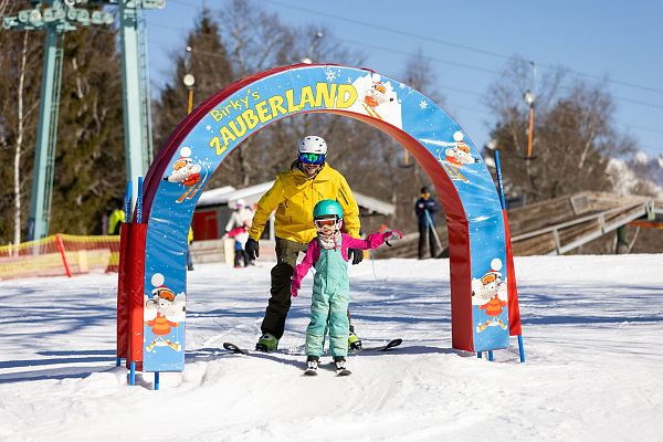 Ein Erwachsener hilft einem Kind beim Skifahren unter einem bunten Tor.