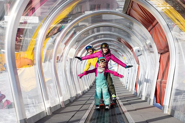Drei Kinder stehen fröhlich auf einem Förderband im Schnee, umgeben von einer transparenten Struktur.