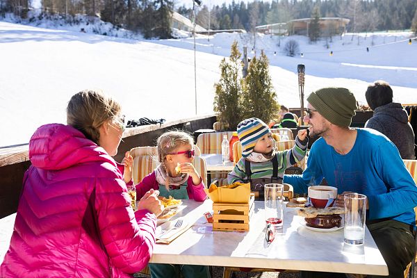Familie sitzt bei Sonnenschein auf einer Terrasse und isst auf einem Tisch mit Blick auf die verschneite Berglandschaft.