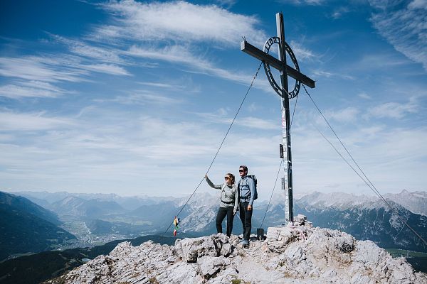 Zwei Personen stehen an einem Berggipfel neben einem großen Kreuz.