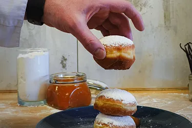 Eine Hand hält einen Krapfen, im Hintergrund Milch und Marmelade.