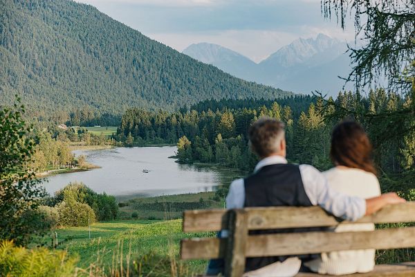 A couple on a bench in front of a lake and mountains, enjoying a peaceful view.