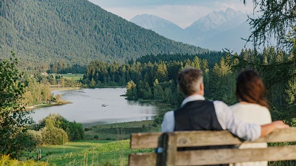 Ein Paar sitzt auf einer Bank und schaut auf einen See, umgeben von Bergen und Bäumen.