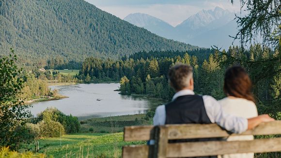A couple on a bench in front of a lake and mountains, enjoying a peaceful view.