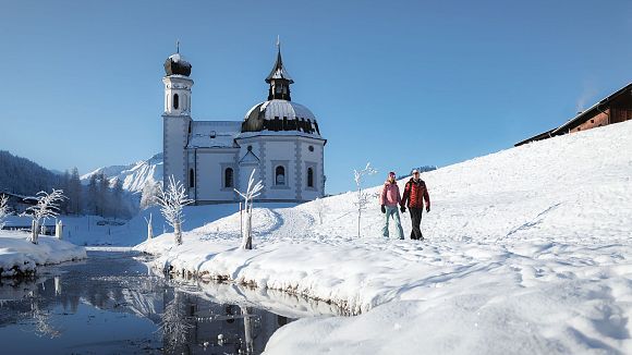Verschneite Kirche in Winterlandschaft, Wanderer im Vordergrund. Region Seefeld. Klarer Himmel, blau.