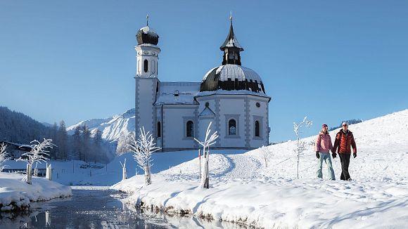 Zwei Wanderer bei einer Winterwanderung vor einer Kirche in einer verschneiten Landschaft.