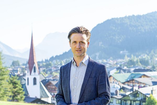 A man in a suit stands with mountains and a village in the background.