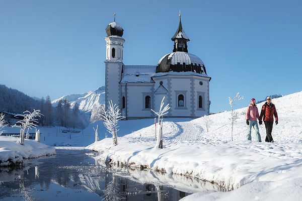 Zwei Wanderer bei einer Winterwanderung vor einer Kirche in einer verschneiten Landschaft.