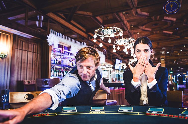 Due persone a un tavolo da gioco in un casinò, un uomo punta e una donna si copre la bocca.