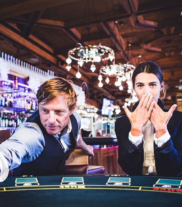 Due persone a un tavolo da gioco in un casinò, un uomo punta e una donna si copre la bocca.
