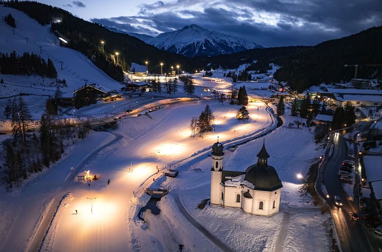A snowy landscape with a church, ski slopes, and mountains, illuminated by evening lights.