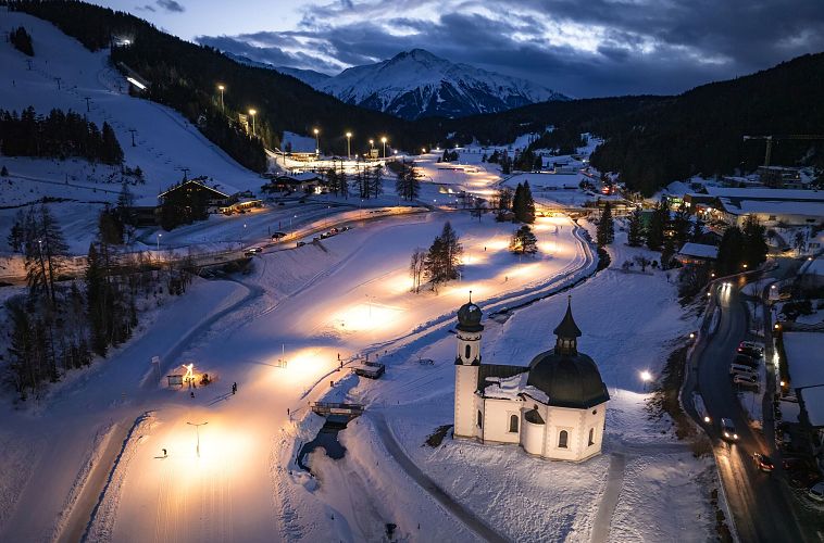 Verschneite Winterlandschaft bei Nacht in der Region Seefeld mit beleuchteter Kirche und Skipisten.