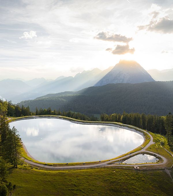 Lago circondato da alberi e montagne sullo sfondo, con il sole che sorge.