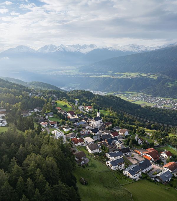 Luftaufnahme eines kleinen Dorfes in bergiger Landschaft mit Hügeln und Bergen im Hintergrund.