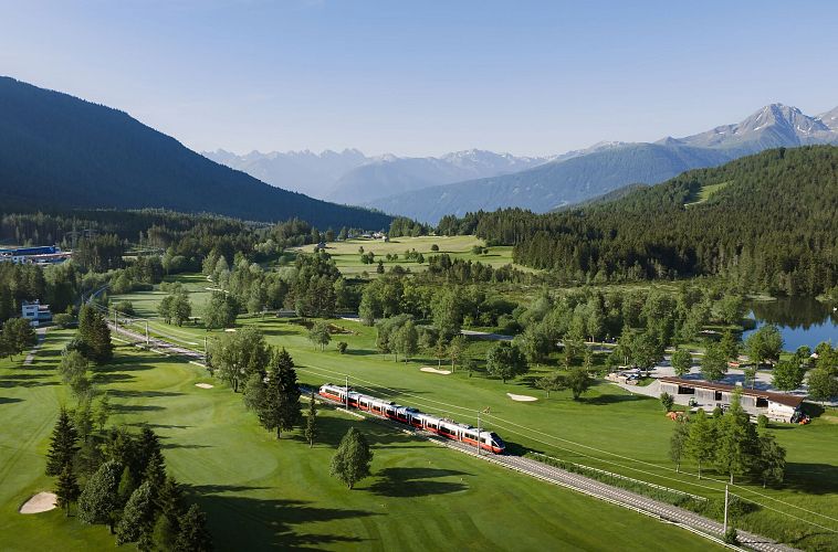 Landschaft mit Golfplatz, Zug und Bergen im Hintergrund.