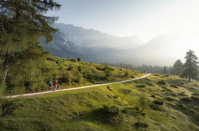 E-Biken im Gaistal im Sommer_Drohnenaufnahme_zwei Biker_innen fahren auf Weg hinauf zur Hämmermoosalm bei Morgenstimmung
