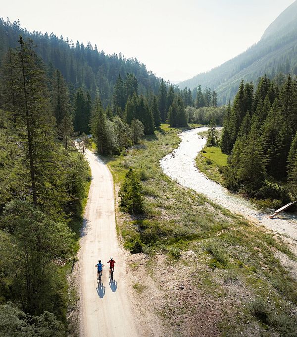 Zwei Radfahrer auf einem Kiesweg entlang eines Flusses in einem bewaldeten Tal.
