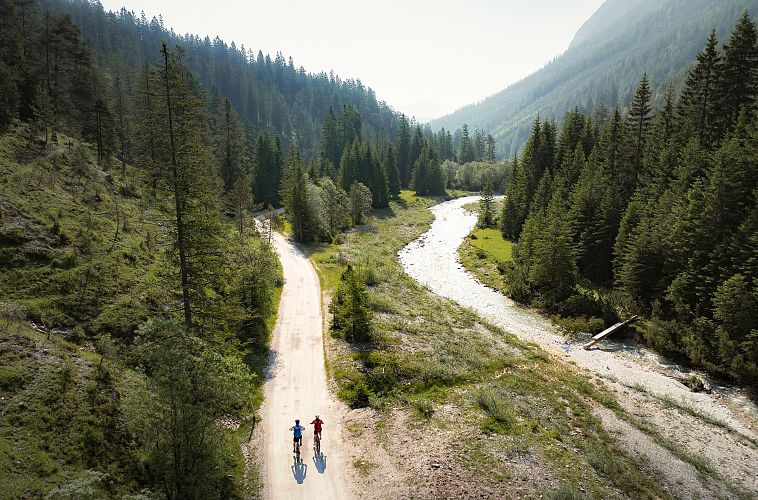 Zwei Radfahrer auf einem Kiesweg entlang eines Flusses in einem bewaldeten Tal.