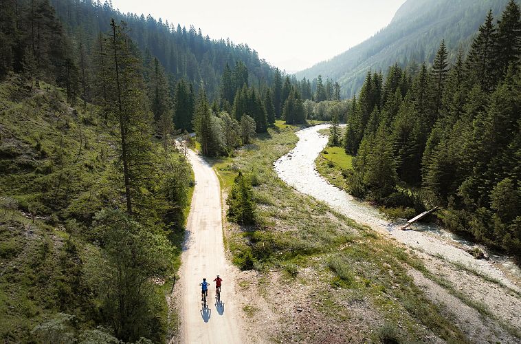 Zwei Radfahrer auf einem Kiesweg entlang eines Flusses in einem bewaldeten Tal.