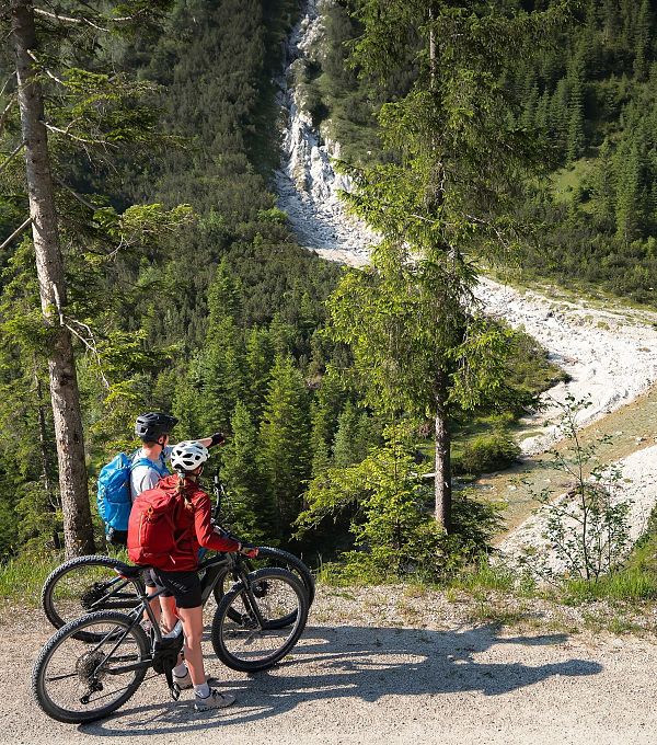 E-Biken im Gaistal im Sommer_zwei Biker_innen auf Forstweg blicken hinab auf Ache