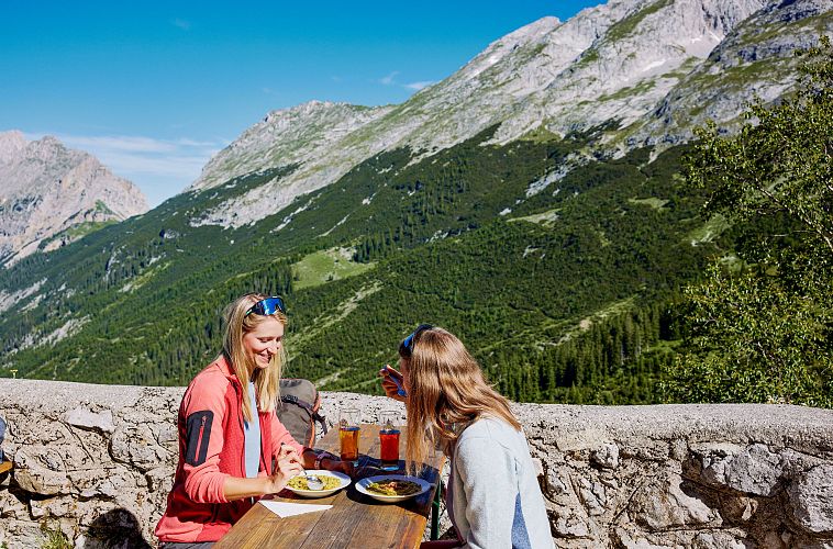 Zwei Frauen sitzen an einem Tisch im Freien und essen, umgeben von Bergen.