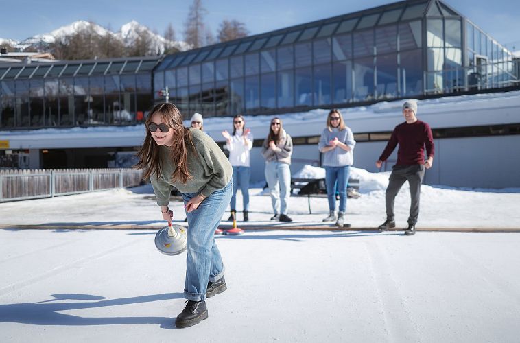 Gruppe spielt Eisstockschießen in der Region Seefeld vor modernem Gebäude auf schneebedeckter Eisfläche.