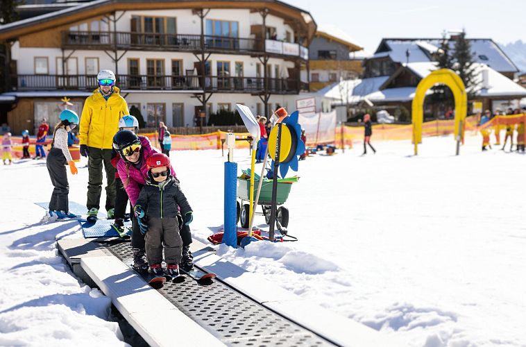 Ein Erwachsener hilft einem Kind beim Skifahren unter einem bunten Tor.