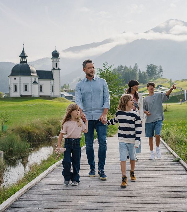 Eine Familie geht auf einem Holzsteg in einer grünen Landschaft mit einer Kirche im Hintergrund.