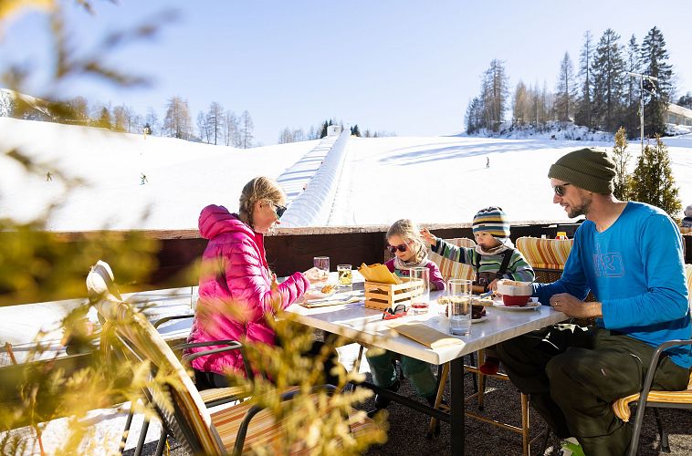 Familie sitzt an einem Tisch auf einer Terrasse und genießt Snacks und Getränke im Schnee.