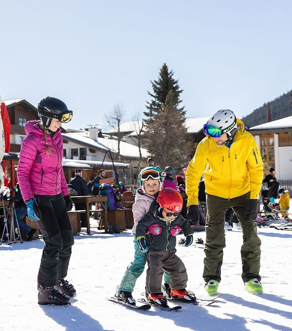 Persone che sciano in una località montana con bambini, in una giornata di sole.