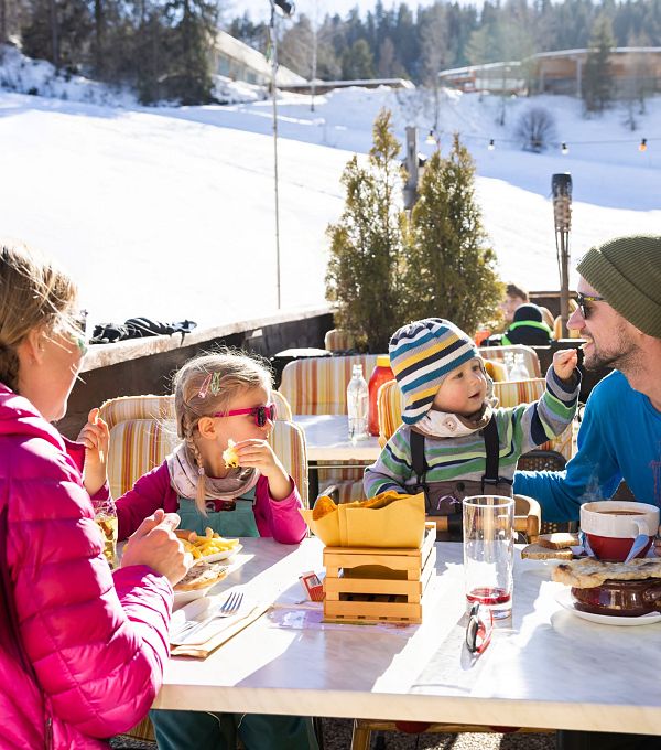 Familie sitzt bei Sonnenschein auf einer Terrasse und isst auf einem Tisch mit Blick auf die verschneite Berglandschaft.