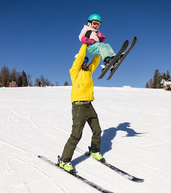 Ein Erwachsener hebt ein Kind beim Skifahren auf einer schneebedeckten Piste.